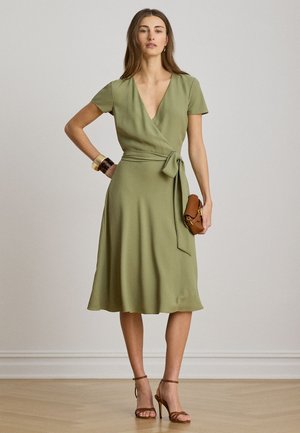 Kvinde i olivengrøn slå-om kjole med korte ærmer, brune højhælede sandaler, armbånd og holder en lille brun clutch.