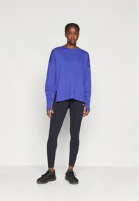 Fioletowa bluzka z długim rękawem z rozcięciami po bokach, zestawiona z czarnymi legginsami. Bluzka ma luźny, swobodny krój i jest wykonana z gładkiej tkaniny. Czarnie sportowe buty dopełniają stylizację.