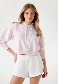 Felpa cropped rosa con logo ricamato, coulisse e maniche a righe, abbinata a pantaloni corti bianchi con vita elastica. Texture liscia, stile casual.