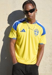 Man with braided hair and sunglasses wearing a yellow and blue Sweden soccer jersey, standing with hands in pockets against a light wall.