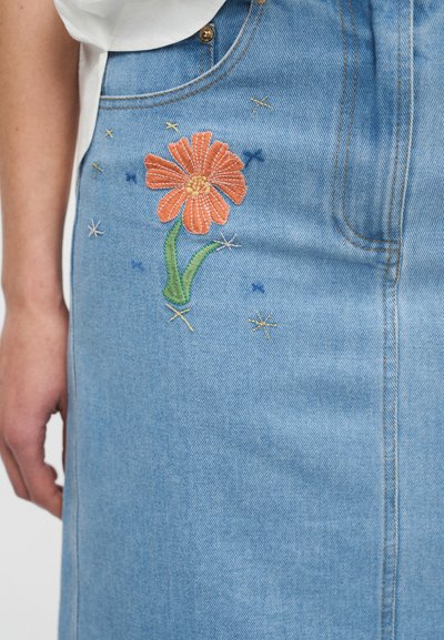 Nærbilde av lys blå denimskjørt med oransje brodert blomst og små dekorative stjernestikninger nær lomma og sidesømmen.