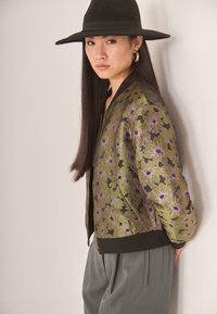 Joven con cabello largo y negro, llevando un sombrero negro de ala ancha, una chaqueta floral verde y púrpura, y pantalones grises, frente a una pared clara.