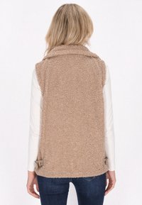 Beige Fleece-Weste mit hohem Kragen, texturiertem Material und seitlichen Kordeldetails. Trägt man über einem langärmeligen weißen Oberteil und kombiniert mit Jeans.