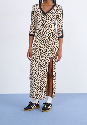 Vrouw draagt een beige midi-jurk met luipaardprint en zwarte bies, een zijsplit, driekwart mouwen, witte sokken en zwarte Adidas-sneakers.