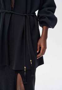 Czarna odzież wierzchnia o teksturowanym wykończeniu, z plecionym paskiem i złotymi akcentami metalowymi. Rękaw jest lekko obszerny, co dodaje wymiaru.