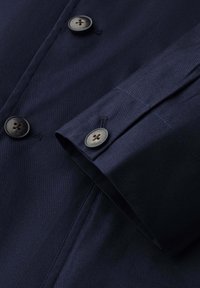 Detailní záběr rukávu a těla tmavě modrého kabátu s tmavými knoflíky s rytinou "Hackett London" a viditelnými stehy.