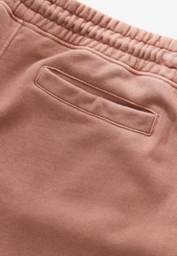 Vue de dos d’un pantalon de survêtement mauve avec une taille élastique et une poche gansée unique avec des coutures propres.