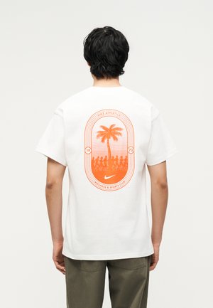 Άτομο που φοράει άσπρο μπλουζάκι με πορτοκαλί γραφικό φοίνικα και δρομείς, με την επιγραφή «Nike Athletics Wellness & Sports Club 1972» στην πλάτη.