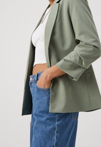 Světle zelený blazer s volným střihem, se tři�čtvrtečními rukávy a hladkou texturou, zkombinovaný s džíny z modré denimu s vysokým pasem.
