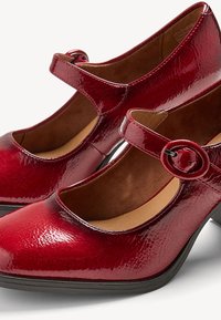 Chaussures Mary Jane rouges brillantes à talons bas, avec des brides à boucle ronde et des semelles intérieures rembourrées marron, présentées côte à côte sur une surface blanche.