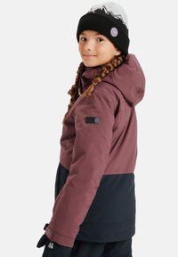 Bordeauxrode en marine winterjas met een capuchon, voorzien van een getextureerde stof en een logo op de mouw. Zwarte beanie met een bergpatroon bovenop.