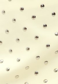 Tissu crème parsemé de clous en argent brillants disposés régulièrement, créant un motif texturé avec une surface lisse dans l'ensemble.