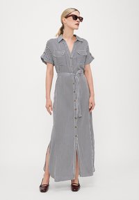 Vestido longo listrado em preto e branco, com mangas curtas dobradas, dois bolsos no peito e cintura ajustada. Fendas laterais e botões dourados.