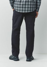 Man wearing dark navy pants, black and gray sneakers, and a blue checkered shirt with hands in pockets, standing on a plain background.