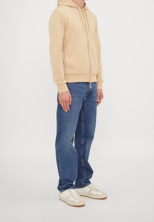 Personne portant un sweat à capuche beige zippé, un jean bleu droit et des baskets blanches, debout devant un fond blanc uni.