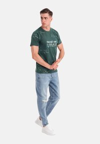 Grøn bomulds T-shirt med hvid tekst, parret med lyseblå slidte jeans og hvide sneakers, der står foran en neutrale baggrund.