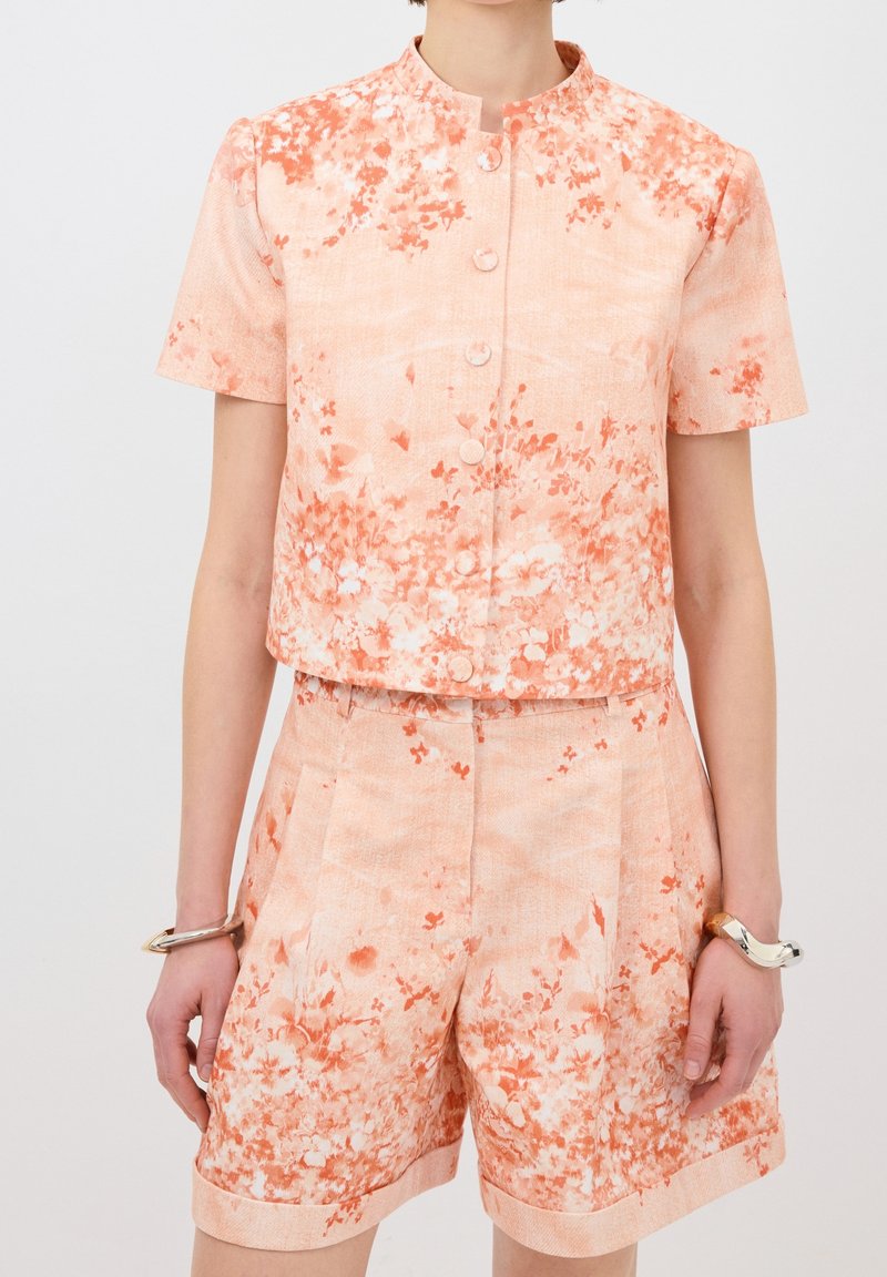 Personne portant un haut à manches courtes à boutons avec un motif floral pêche et orange, assorti à un short taille haute, ainsi que des bracelets argentés aux deux poignets.