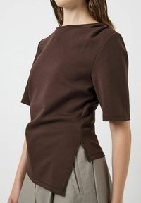 Maglietta marrone a maniche corte con scollo rotondo, texture liscia, orlo asimmetrico con spacchi laterali, abbinata a pantaloni alti beige chiaro.