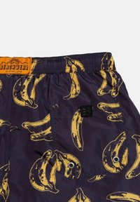 Pantaloni da nuoto con tessuto viola e motivo a banana giallo. Vita elastica con etichetta arancione "BEACHWEAR" e hardware nero.