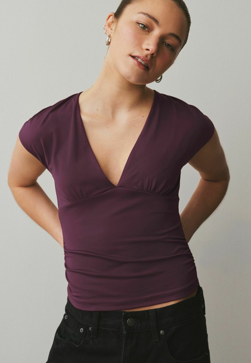 Jeune femme aux cheveux attachés, portant un haut col en V violet foncé et un pantalon noir, debout les mains derrière le dos devant un fond uni.