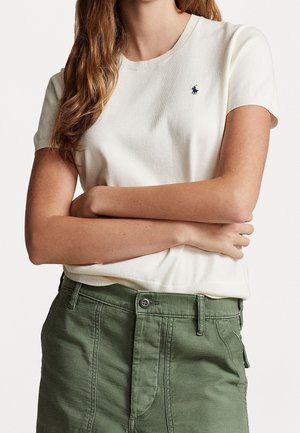 Vrouw met gekruiste armen, gekleed in een wit T-shirt met korte mouwen met een klein marineblauw logo en een groen broek met hoge taille, knoop en riemlussen.