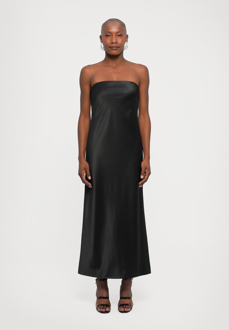 Vestido maxi de satin preto, sem alças, com um decote reto e uma silhueta elegante. O modelo usa sandálias pretas com tiras.