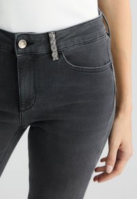 Pelēki denim džinsi ar šauru piegriezumu, ar dekoratīvu rhinestone akcentu pogu aizdarei un priekšējām kabatām. Gluda tekstūra.