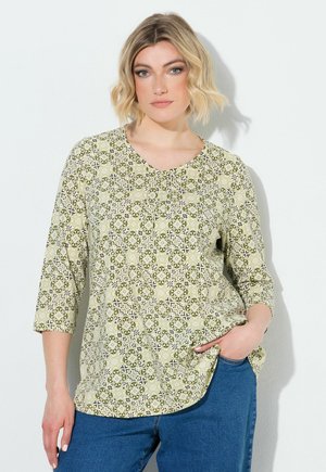 Donna con capelli biondi corti che indossa una maglia a maniche tre quarti verde chiaro a motivi e jeans blu, in piedi davanti a uno sfondo bianco.