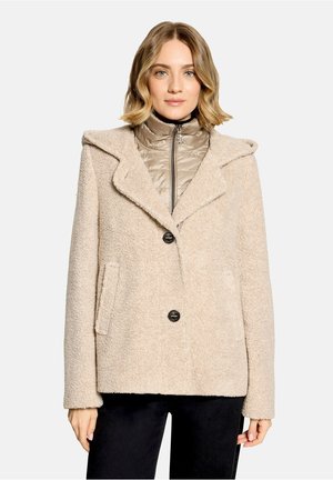 Femme portant un manteau teddy beige avec deux boutons sur une veste matelassée zippée, associé à un pantalon noir, debout devant un fond uni.