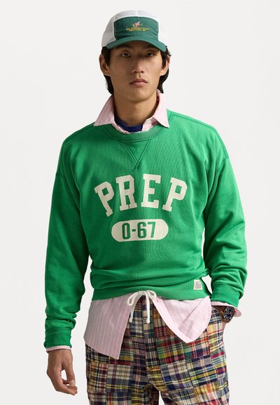 Green sweatshirt with "PREP 0-67" printed in off-white, layered over a pink striped shirt, worn with multicolored plaid shorts and a cap.