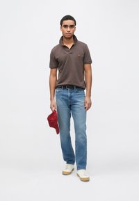 Polo castanha com um pequeno logótipo, jeans azuis, ténis creme, segurando um boné de beisebol vermelho, em pé contra um fundo branco.