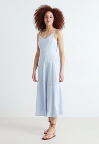 Rochie midi fără mânecă, de un albastru deschis, cu decolteu în V, dintr-un material texturat și detalii cu ochiuri; purtată cu sandale maronii cu barete.