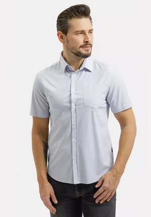 Młody mężczyzna z brodą, ubrany w jasnoniebieską koszulę na krótki rękaw z guzikami i ciemne spodnie, patrzący na bok na jednolitym tle.