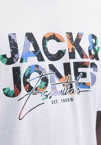 Balts kokvilnas T-krekls ar lieliem melniem uzrakstiem "JACK & JONES Essentials" un krāsainiem traipu akcentiem zaļā, zilā, oranžā un purpura krāsā.