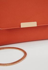 Gros plan d’un sac à main en cuir orange avec un fermoir rectangulaire doré et une chaîne dorée tressée sur une surface blanche.