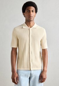 Cream short-sleeve knit polo shirt with textured open weave, button-down front, and contrasting collar. Paired with light denim jeans.
