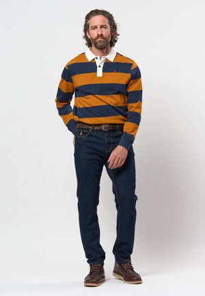Mann mit Bart und welligem Haar, der ein langärmeliges Polohemd mit senf- und marineblauen Streifen, dunkle Jeans, braunen Gürtel und braune Stiefel trägt, steht.