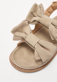 Beige mokkanahka sandaalit, joissa on kaksi suurta kaarimittaria, avoimet sivut ja kirjailtu ruskea pohja. Pehmeä rakenne ja litteä muotoilu.