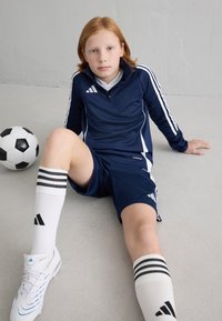 Fato de treino atlético azul-marinho com acentos brancos, combinado com meias brancas com riscas pretas e sapatilhas brancas. Uma bola de futebol em preto e branco próxima.