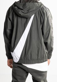 Mörkgrön huva jacka i lätt material, med ett stort vitt diagonalt rand och elastiska manschetter vid midjan och ärmarna.