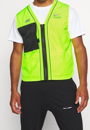 Mężczyzna ubrany w neonowozieloną kamizelkę Nike z przednim zamkiem błyskawicznym, na białej koszulce i czarnych spodniach z napisem "Night Addict" na udzie.