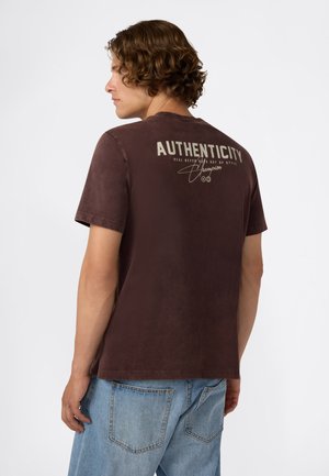 Jonge man met krullend haar, gekleed in een bruine T-shirt met de tekst "AUTHENTICITY" en lichtblauwe jeans, die naar links kijkt op een witte achtergrond.