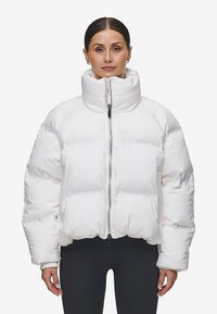 Vit pufferjacka med hög krage, dragkedjestängning och raglanärmar. Den har en quiltad design med synliga sömmar och elastiska ärmslut.