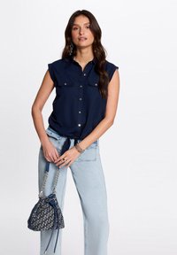 Blouse sans manches bleu marine avec un devant boutonné et deux poches poitrine, assortie à un jean taille haute bleu clair et un sac seau à motifs.