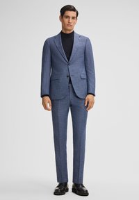 Abito a quadri blu con blazer aderente e pantaloni, realizzato in tessuto testurizzato. Collo alto nero indossato sotto e scarpe nere.