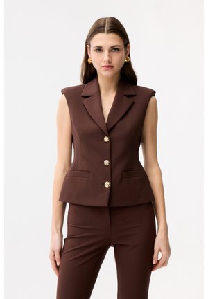 Kobieta o prostych brązowych włosach nosi bezrękawnikowy brązowy garnitur z złotymi guzikami i dopasowane spodnie, stojąc na tle jednolitego białego tła.