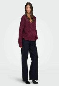 Bordeauxfarbener strukturierter Pullover mit V-Ausschnitt, kombiniert mit dunkelblauen weiten Jeans und schwarzen High Heels.