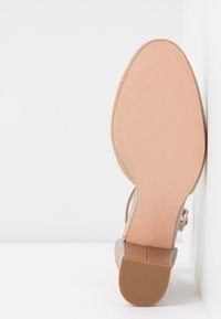 Sandale à talon marron avec une semelle extérieure beige clair, présentant une texture lisse, un bout arrondi et une fine sangle avec des accents métalliques.