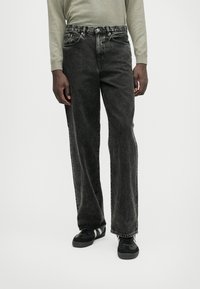 Črne jean hlače s širokim krojem, z nenatančnim zaključkom, petimi žepi in kovinskim gumbom za zapiranje. Uporabljene z črnimi supergami.