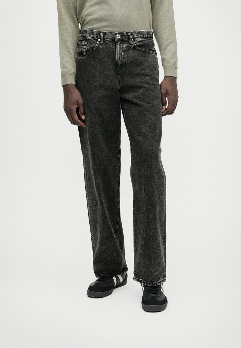 Črne jean hlače s širokim krojem, z nenatančnim zaključkom, petimi žepi in kovinskim gumbom za zapiranje. Uporabljene z črnimi supergami.